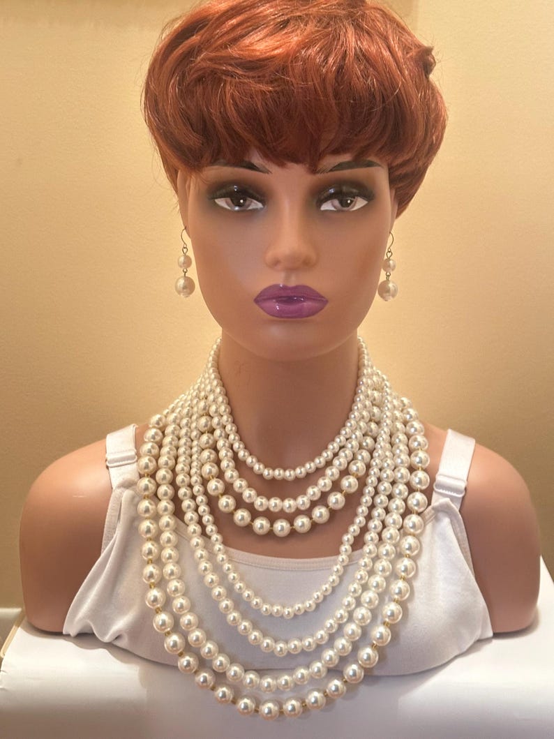 May include: A mannequin head with a red wig and pearl jewellery. The jewellery includes a set of pearl earrings and multiple strands of white faux pearl necklaces. The mannequin is wearing a white vest top.