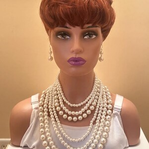 May include: A mannequin head with a red wig, pearl earrings, and a multi-strand pearl necklace. The necklace features several layers of varying pearl sizes. The mannequin is wearing a white vest top.