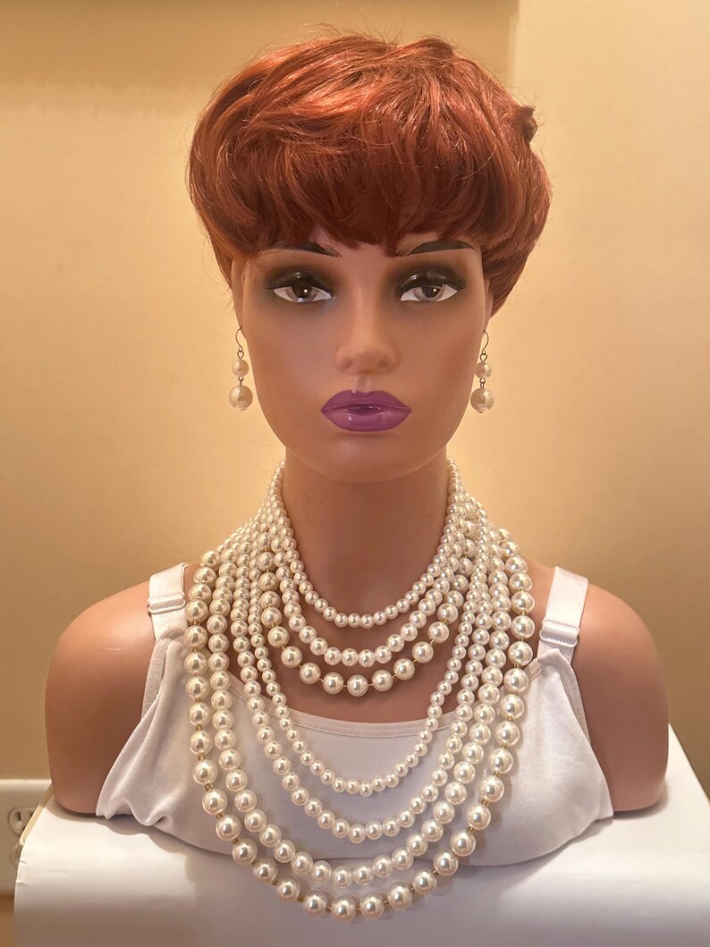 May include: A mannequin head with a red wig, pearl earrings, and a multi-strand pearl necklace. The necklace features several layers of varying pearl sizes. The mannequin is wearing a white vest top.