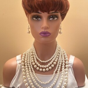 May include: A mannequin head with a red wig, adorned with a multi-strand pearl necklace and matching pearl earrings. The mannequin is also wearing a white vest top. The background is a plain, light beige colour.