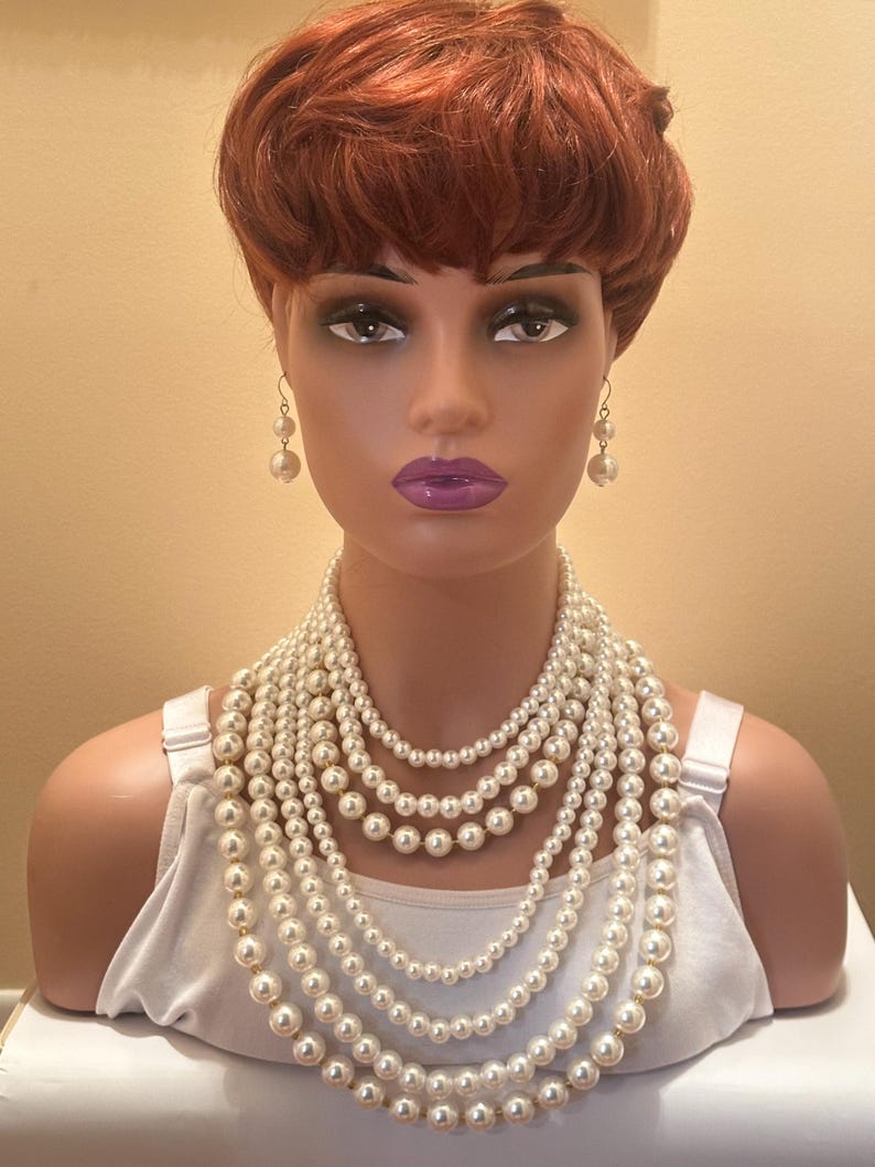 May include: A mannequin head with a red wig, adorned with a multi-strand pearl necklace and matching pearl earrings. The mannequin is also wearing a white vest top. The background is a plain, light beige colour.