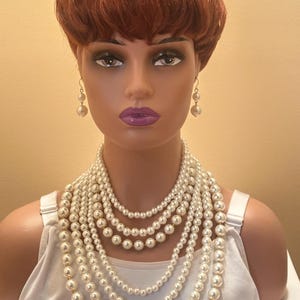 May include: A mannequin head with a red wig displays a multi-strand pearl necklace and matching pearl earrings. The necklace features varying sizes of cream-coloured pearls, creating a layered effect. The mannequin wears a white vest top.