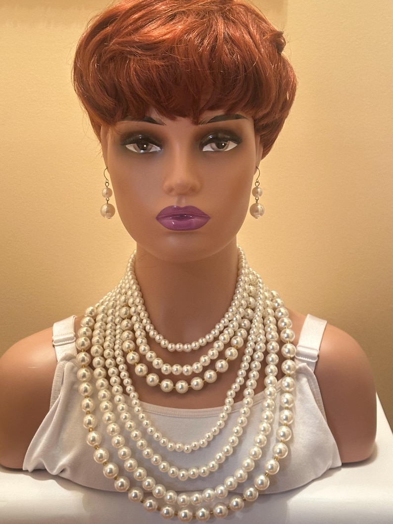 May include: A mannequin head with a red wig displays a multi-strand pearl necklace and matching pearl earrings. The necklace features varying sizes of cream-coloured pearls, creating a layered effect. The mannequin wears a white vest top.