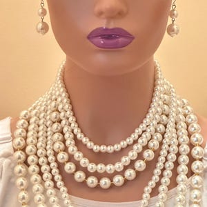 May include: A mannequin wearing a multi-strand pearl necklace and matching pearl earrings. The necklace features varying sizes of white pearls. The earrings are a drop style with two pearls. The mannequin has purple lipstick.