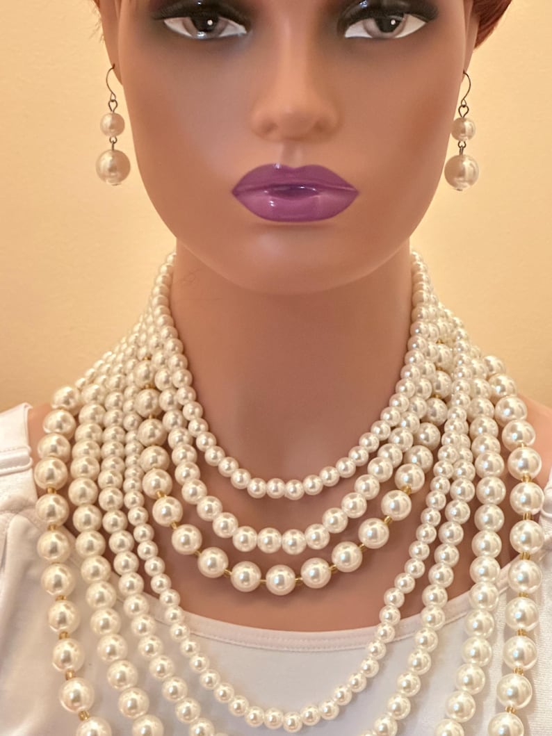 May include: A mannequin wearing a multi-strand pearl necklace and matching pearl earrings. The necklace features varying sizes of white pearls. The earrings are a drop style with two pearls. The mannequin has purple lipstick.