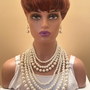 May include: A mannequin head adorned with a multi-strand pearl necklace and pearl drop earrings. The necklace comprises several layers of varying lengths, creating a cascading effect. The pearls are a creamy white hue, and the mannequin has red hair and purple lipstick.