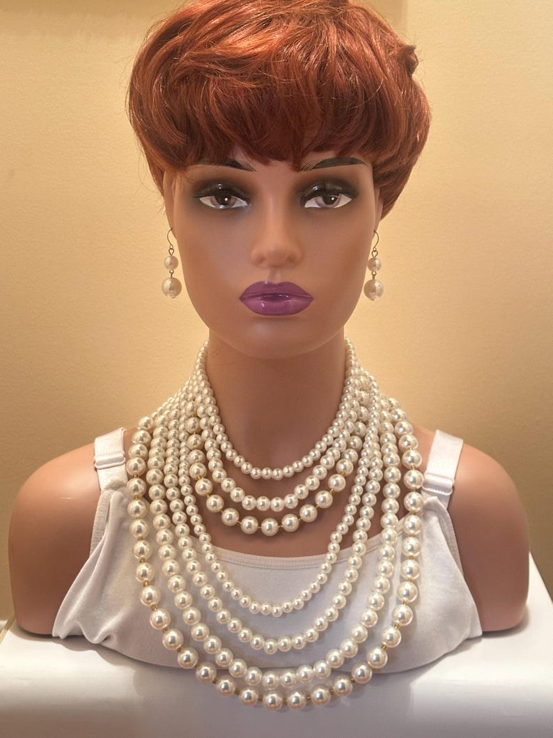 May include: A mannequin head adorned with a multi-strand pearl necklace and pearl drop earrings. The necklace comprises several layers of varying lengths, creating a cascading effect. The pearls are a creamy white hue, and the mannequin has red hair and purple lipstick.