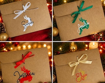 May include: Four rectangular brown kraft paper envelopes, each tied with a satin ribbon bow. Each envelope features a unique vinyl decal of a leaping reindeer silhouette paired with a custom name in script font: Sarah, Amelia, Jessica, and Elyse.