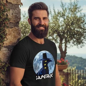 May include: Black t-shirt featuring a graphic of a figure in a top hat against a moonlit background, with the word "HUNTRAX" below. The shirt is worn by a person in blue jeans.