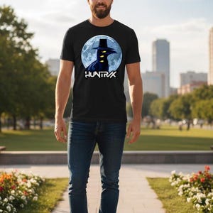 May include: Black t-shirt featuring a graphic of a figure in a hat against a full moon, with the word "HUNTRX" below. The shirt is paired with blue jeans and white sneakers. The person is standing on a paved walkway in a park.
