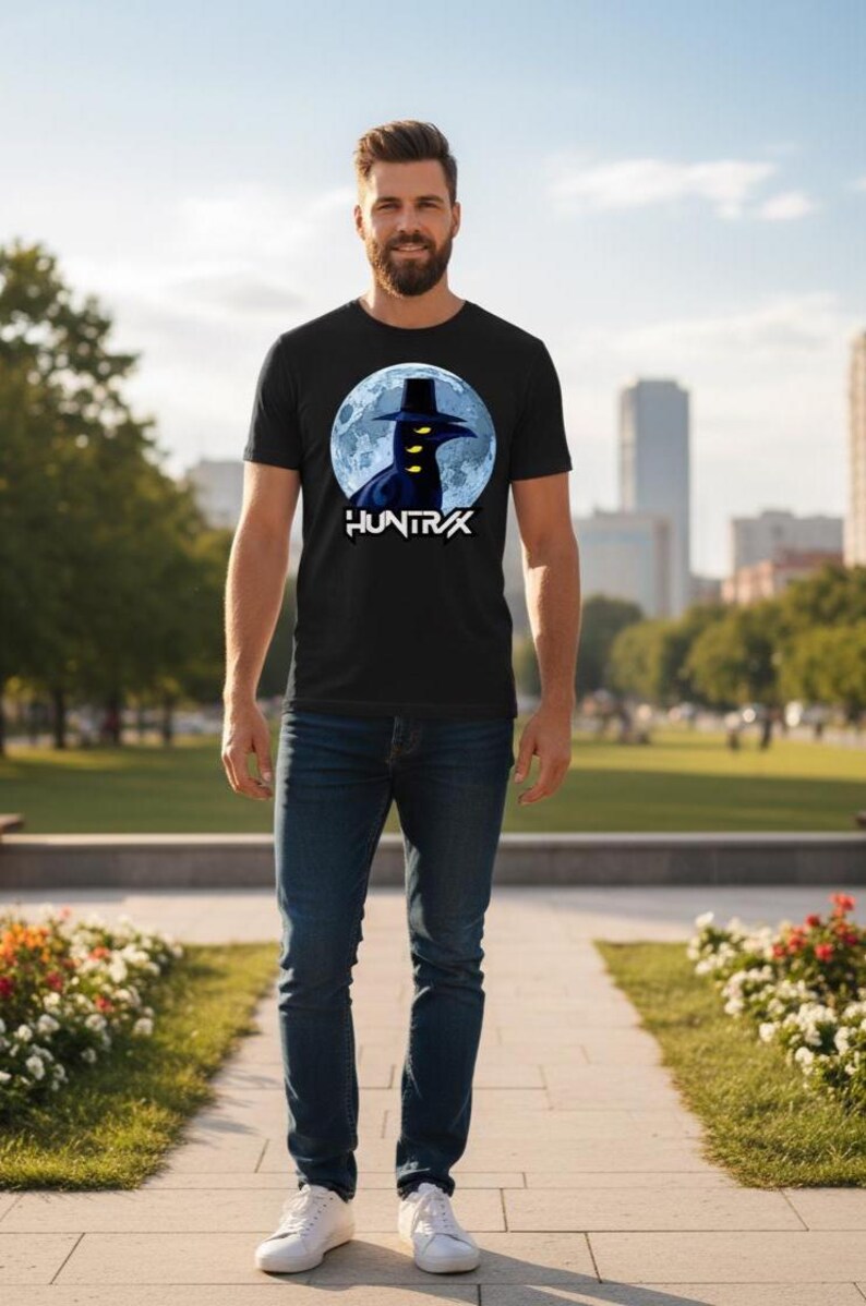 May include: Black t-shirt featuring a graphic of a figure in a hat against a full moon, with the word "HUNTRX" below. The shirt is paired with blue jeans and white sneakers. The person is standing on a paved walkway in a park.