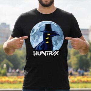 May include: Black t-shirt featuring a graphic of a dark figure with a hat and yellow eyes against a blue moon backdrop. The word "HUNTRX" is printed in white below the graphic. The shirt is a crew neck.