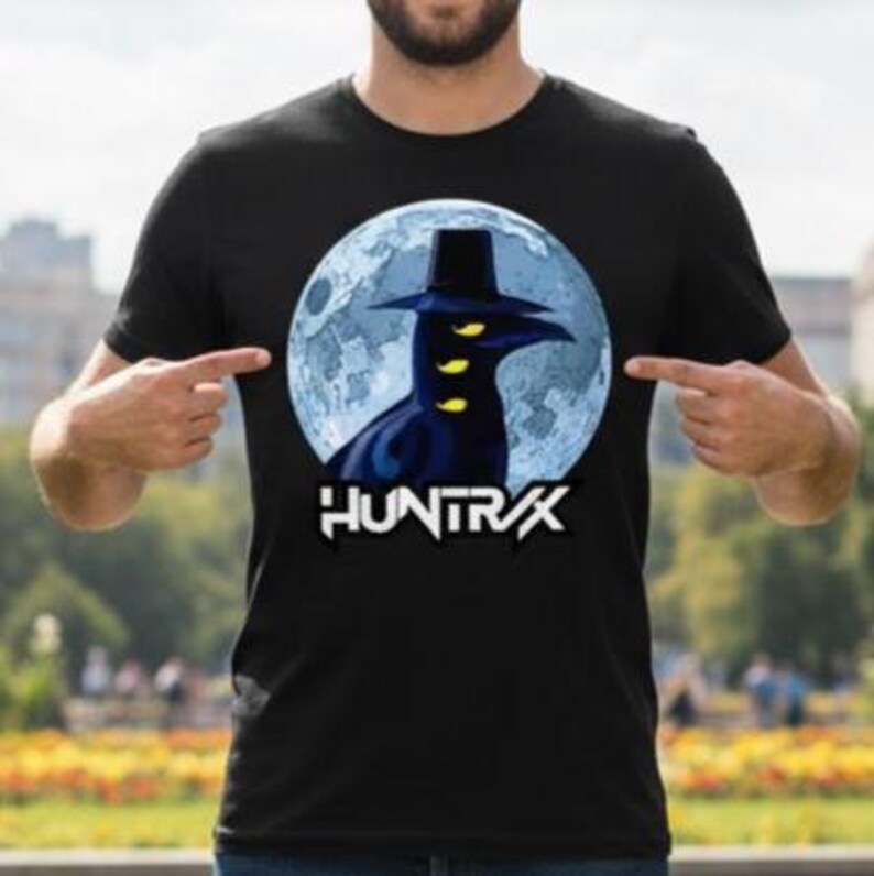 May include: Black t-shirt featuring a graphic of a dark figure with a hat and yellow eyes against a blue moon backdrop. The word "HUNTRX" is printed in white below the graphic. The shirt is a crew neck.
