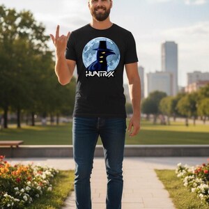 May include: Black t-shirt featuring a graphic of a crow wearing a hat in front of a full moon, with the word "HUNTRAX" below. The person is wearing blue jeans and white sneakers.