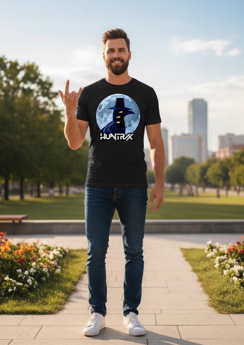 May include: Black t-shirt featuring a graphic of a crow wearing a hat in front of a full moon, with the word "HUNTRAX" below. The person is wearing blue jeans and white sneakers.