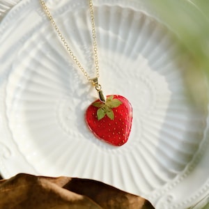 May include: A gold chain necklace with a red strawberry pendant. The strawberry is made of resin and has a dried flower inside.