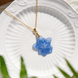 Peut inclure: Un collier délicat en chaîne dorée avec un pendentif en forme de fleur bleue. La fleur est faite de fleurs réelles pressées et est enfermée dans de la résine. Le collier est un bijou magnifique et unique.