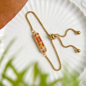 May include: Gold-toned adjustable bracelet featuring a rectangular charm with a red background and embedded dried yellow and white flowers. The bracelet has a delicate chain and a sliding bead for adjustment.