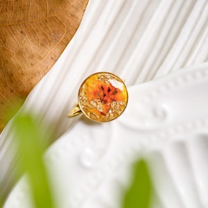 Könnte beinhalten: Goldener Ring mit einem großen, runden, harzgefüllten Einsatz. Der Einsatz enthält eine getrocknete orangefarbene Blume und Goldflocken. Der Ring liegt auf einer weißen Oberfläche.