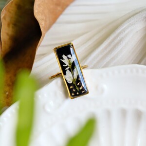 Puede incluir: Un anillo dorado con un engaste rectangular de resina negra. El engaste presenta una delicada flor blanca y copos dorados. El anillo se muestra sobre una superficie blanca, resaltando el diseño floral.
