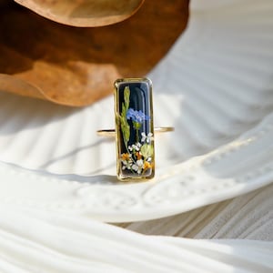 May include: A gold ring with a rectangular black resin setting featuring pressed flowers in shades of blue, white, and yellow.
