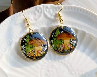 Op de afbeelding: Een paar gouden paddenstoel oorbellen met geperste bloemen en een donkere achtergrond. De oorbellen zijn rond en hebben een gouden haak.