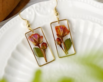 May include: A pair of gold-colored rectangular earrings. Each earring features a dried orange and pink flower encased in clear resin. The earrings have gold-colored hooks and are displayed on a white surface.