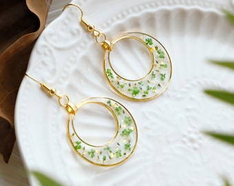 Pode incluir: Um par de brincos de argola dourados. Cada brinco apresenta um círculo de resina transparente preenchido com pequenos trevos verdes e flocos de ouro. Os brincos são exibidos em um prato branco.
