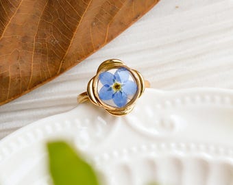 May include: A gold ring with a blue forget-me-not flower encased in resin. The flower is visible through the clear resin.