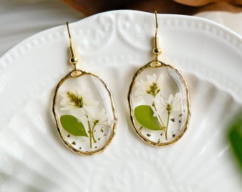 May include: A pair of gold oval earrings with white flowers and green leaves pressed inside clear resin.