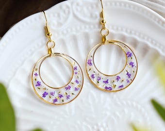 May include: A pair of gold hoop earrings with a clear resin design featuring small purple pressed flowers.