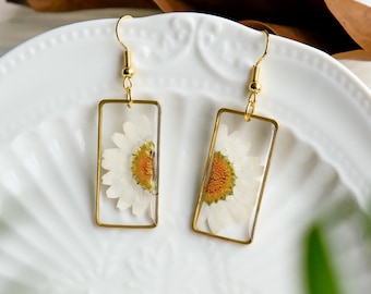 May include: A pair of gold earrings with a rectangular frame and a pressed white daisy flower inside each frame.