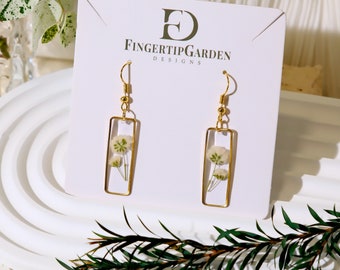 May include: Gold earrings with a rectangular frame and a small white flower pressed inside. The earrings are on a white card with the text "FINGER TIP GARDEN DESIGNS".
