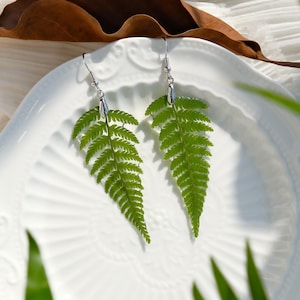 Könnte beinhalten: Ein Paar silberne Ohrringe mit grünen Farnblatt-Anhängern. Die Ohrringe befinden sich auf einem weißen Hintergrund.