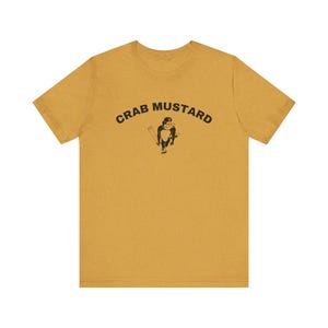 May include: Mustard yellow t-shirt with the text "CRAB MUSTARD" above a black cartoon illustration of a crab holding a baseball bat. The shirt is made of a soft material and has a crew neck.