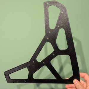 May include: A black, geometric metal bracket with a textured surface. The bracket has an open, angular design with multiple holes and cutouts. It is held against a green background.