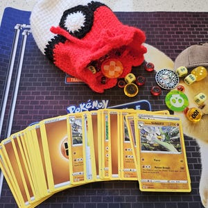 Puede incluir: Una bolsa de ganchillo roja, blanca y negra, que se asemeja a una Poké Ball, está abierta junto a una baraja de cartas Pokémon amarillas. Dados, fichas y un pequeño sombrero están esparcidos alrededor de las cartas sobre un tapete de juego.