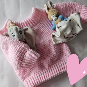 May include: A pale pink, hand-crocheted baby sweater with a ribbed collar and cuffs. A gray stuffed elephant and a brown stuffed bunny are nestled in the sweater. The bunny is wrapped in a white blanket with a blue and grey pattern. A pink heart is in the corner.