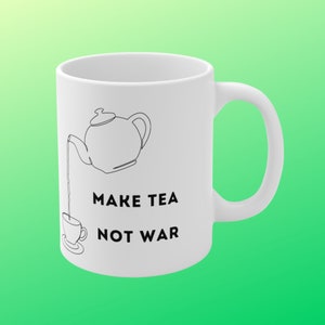 Pode incluir: Caneca de cerâmica branca com um desenho de linha preta de um bule despejando chá em uma xícara. O texto "MAKE TEA NOT WAR" está impresso abaixo da imagem.