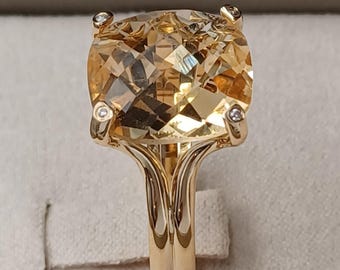 Pode incluir: Um anel de ouro com uma grande pedra preciosa castanho-clara com corte almofada. O anel tem um design de haste dividida e pequenos diamantes de destaque em cada canto da pedra preciosa. O anel está sobre um fundo neutro.