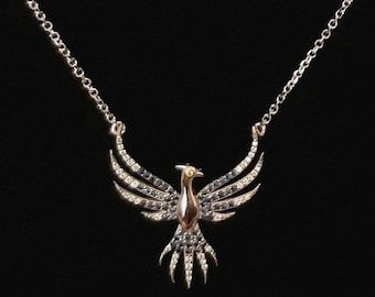 May include: A silver necklace with a pendant shaped like a phoenix in flight. The bird has a gold-colored body, with wings and tail detailed with clear and dark gemstones. The necklace chain is also silver, against a black background.
