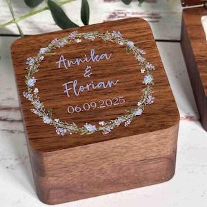 Könnte beinhalten: Eine Holz-Ringbox mit floralem Kranzdesign. Die Box ist mit den Namen "Annika & Florian" und dem Datum "06.09.2025" in hellem Lila graviert. Der Kranz zeigt kleine blaue Blüten und grüne Blätter.