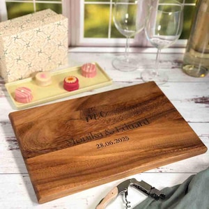 Könnte beinhalten: Rechteckiges Holzschneidebrett mit eingraviertem Text "Mr & Mrs Monika & Eduard 28.06.2025". Daneben ein Korkenzieher, ein Tablett mit Desserts, Weingläser und eine Geschenkbox.