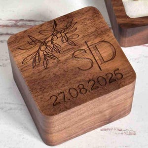 Könnte beinhalten: Eine quadratische Holzbox mit Scharnierdeckel. Die Box ist mit Olivenzweigdetails, den Initialen "S/D" und dem Datum "27.08.2025" graviert. Das Holz hat einen warmen Braunton, und die Gravur ist dunkel.