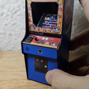 Op de afbeelding: Een miniatuurreplica van een Donkey Kong-arcadekast met een blauw frame, een scherm met spelafbeeldingen, een rode joystick en een bedieningspaneel met karakterillustraties. De bovenkant toont het Donkey Kong-logo in blauwe tekst.