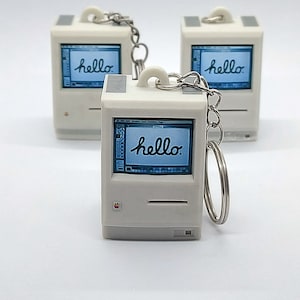 May include: Three white keychain replicas of an old Apple computer with a blue screen that says "hello" on it.