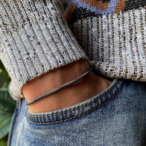 May include: A black braided bracelet on a person's wrist. The bracelet is made of a thin, braided cord and is worn over a gray sweater and blue jeans.
