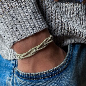 May include: A light beige braided bracelet. The bracelet is made from multiple woven strands, giving it a textured look. It is worn on a wrist, with a grey sweater and blue jeans visible in the background.