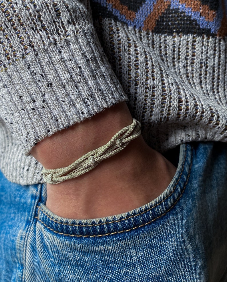 May include: A light beige braided bracelet. The bracelet is made from multiple woven strands, giving it a textured look. It is worn on a wrist, with a grey sweater and blue jeans visible in the background.