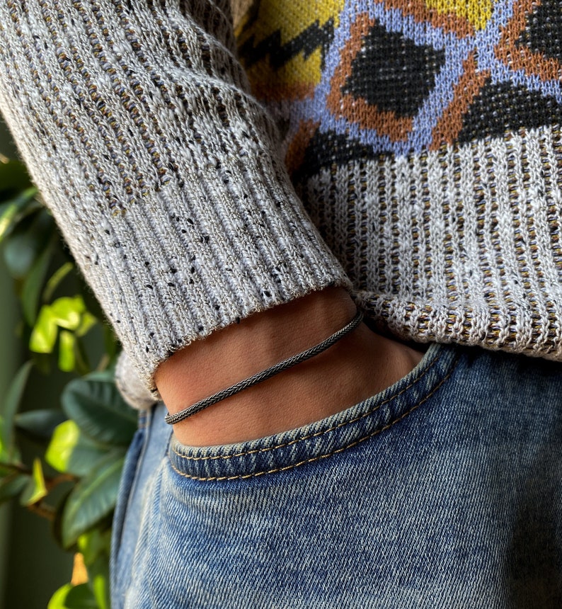 May include: A black braided bracelet on a person's wrist. The bracelet is made of a thin, braided cord and is worn over a gray sweater and blue jeans.